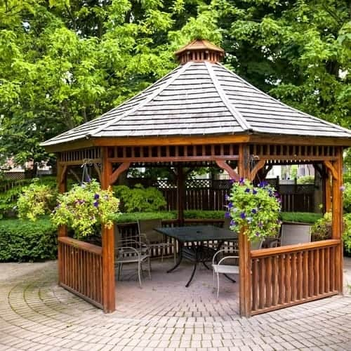 Brown wooden gazebo surrounded by hanging potted plants with bushes and trees on stone bricks