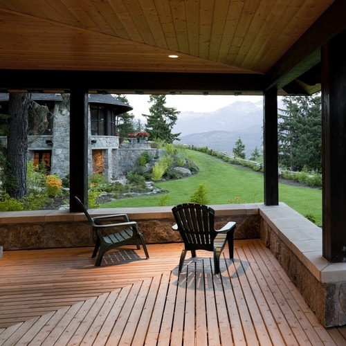 Outdoor sitting area with brown wooden panel flooring and ceiling with black pillars with gray stone house with bushes and trees in background with distant mountains on cloudy day