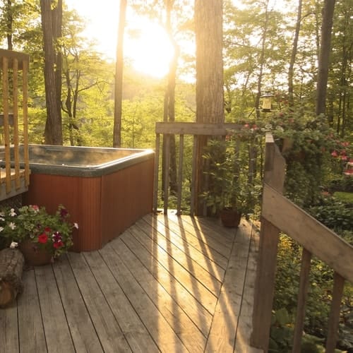 Outdoor brown wooden deck that's fenced in with hot tub and plants around it surrounded by trees and sunlight shining through onto the deck