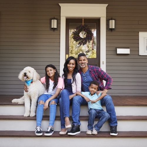 Man, woman, and two children with white dog on white and brown wooden steps on outdoor porch on house with greenish-brown paneled walls with two lights, brown door, and wreath 