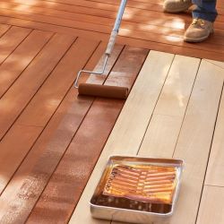 Person with boots with extended paint roller painting brown wood stain on light tan wooden panels with metal paint holder on ground