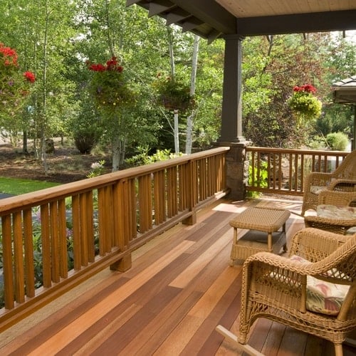 Outdoor brown fenced-in wooden deck with tan wooden chairs with potted plants hanging from roof surrounded by trees