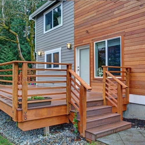 Gray and brown split wooden paneled wall with brown wooden outdoor deck with steps leading to stone path with trees in background 